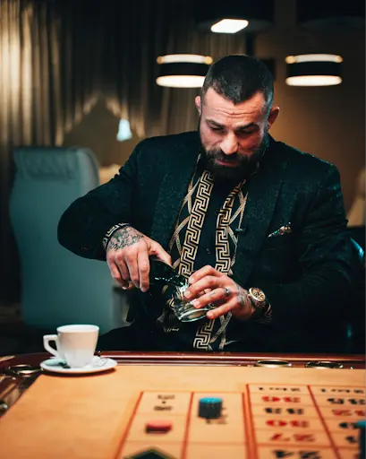 Stylový hráč u ruletového stolu s drinkem v ruce. Muž v luxusním saku s tetováním na rukou nalévá alkohol do sklenice u ruletového stolu, vedle šálku kávy.